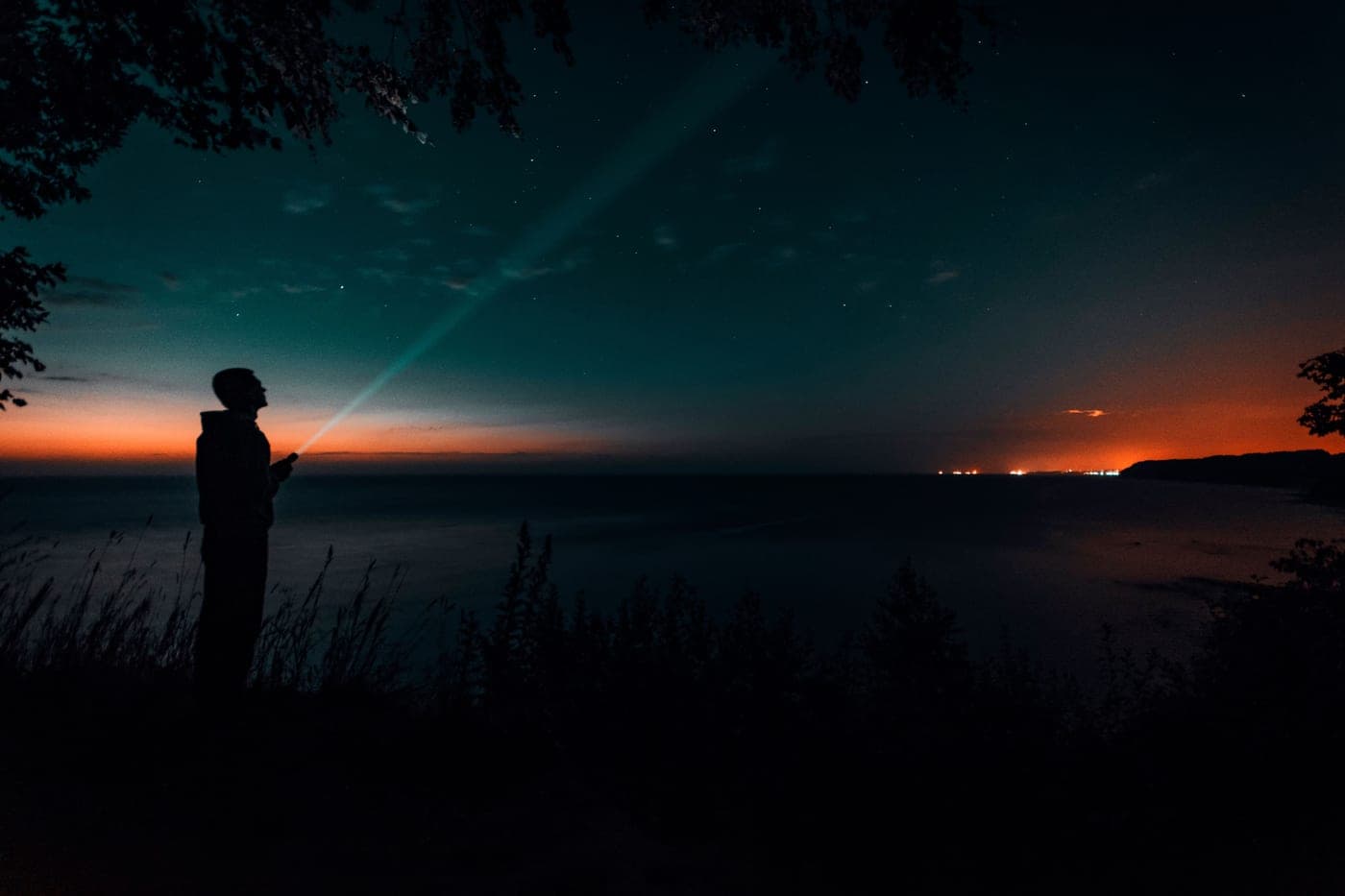 A person with a flashlight silhouetted against a starry twilight sky by the sea
