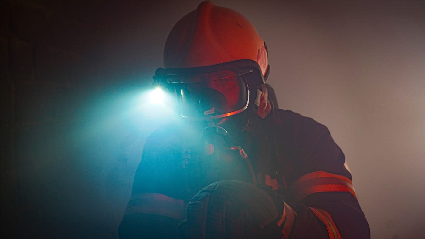 A firefighter in protective gear using a flashlight in heavy smoke