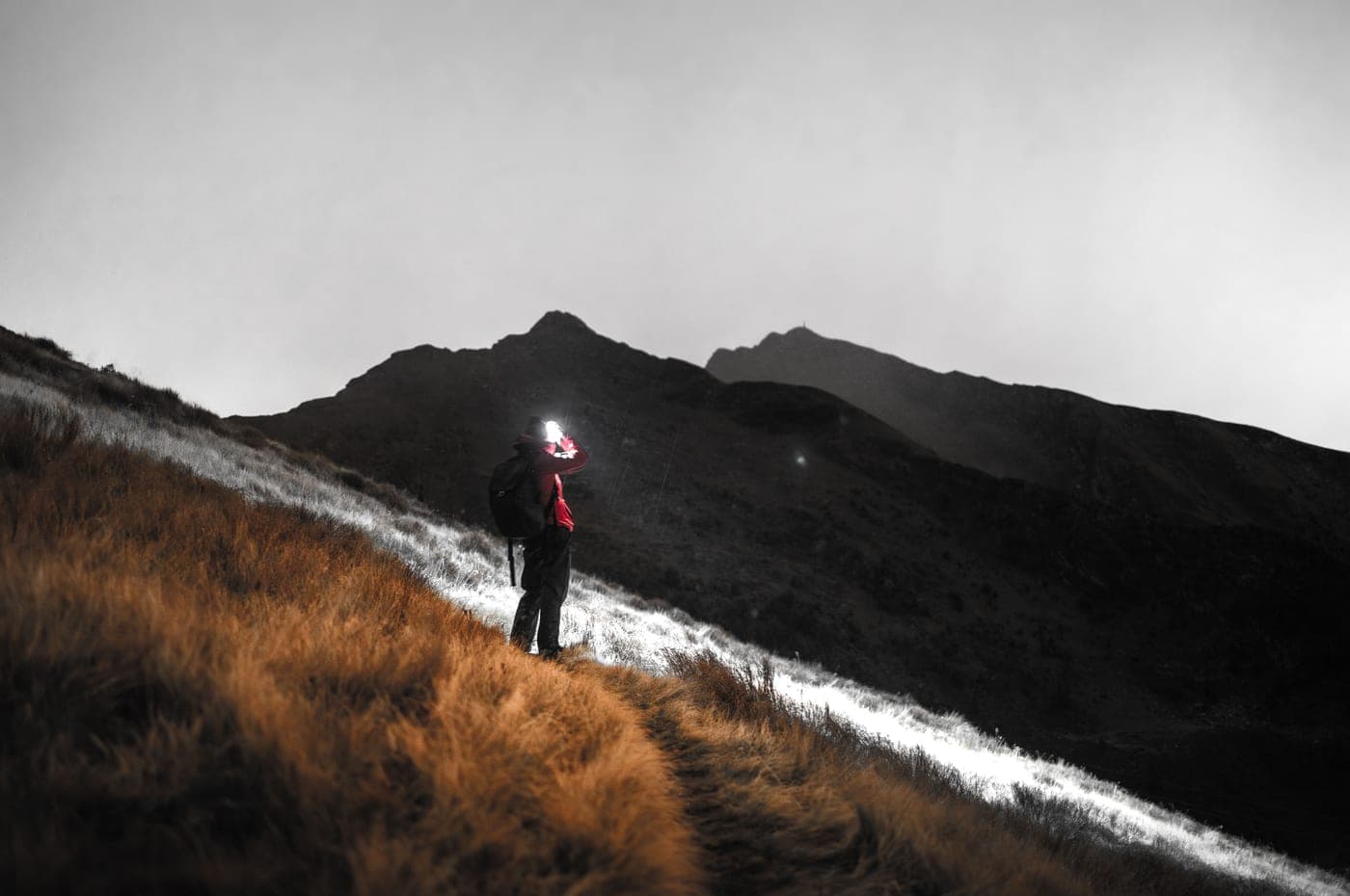 A lone hiker lit by a headlamp on a mountain path under a cloudy sky