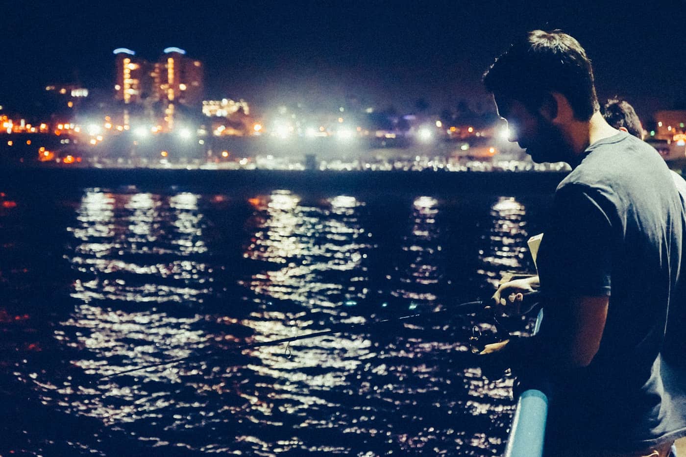 A person fishing from a pier at night with lights reflected on the water