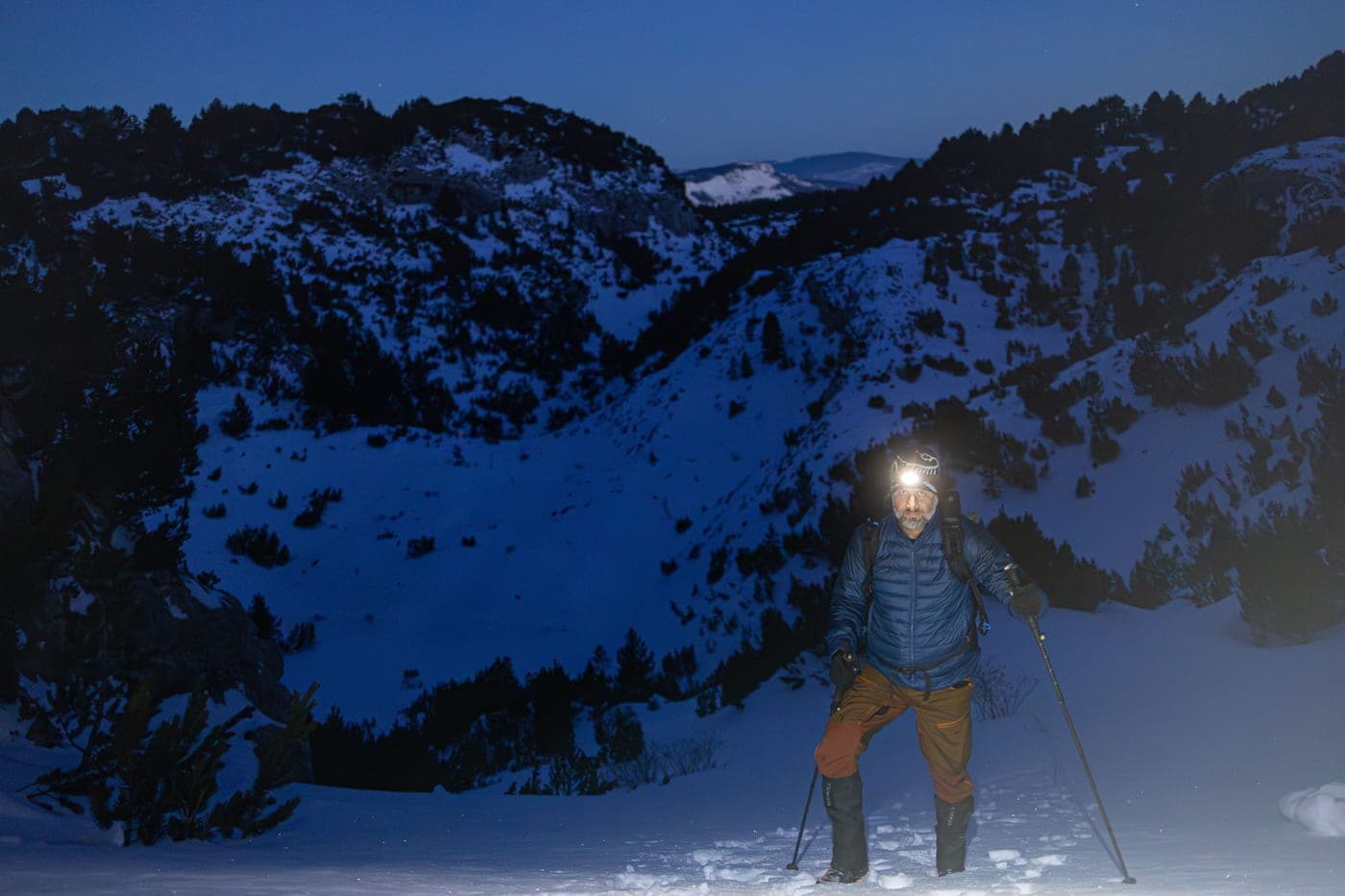 A lone hiker climbing a snowy mountainside at night by headlamp